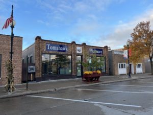 A brick building labeled as Tomahawk Pharmacy with blue sky background and an American flag waving on a flagpole
