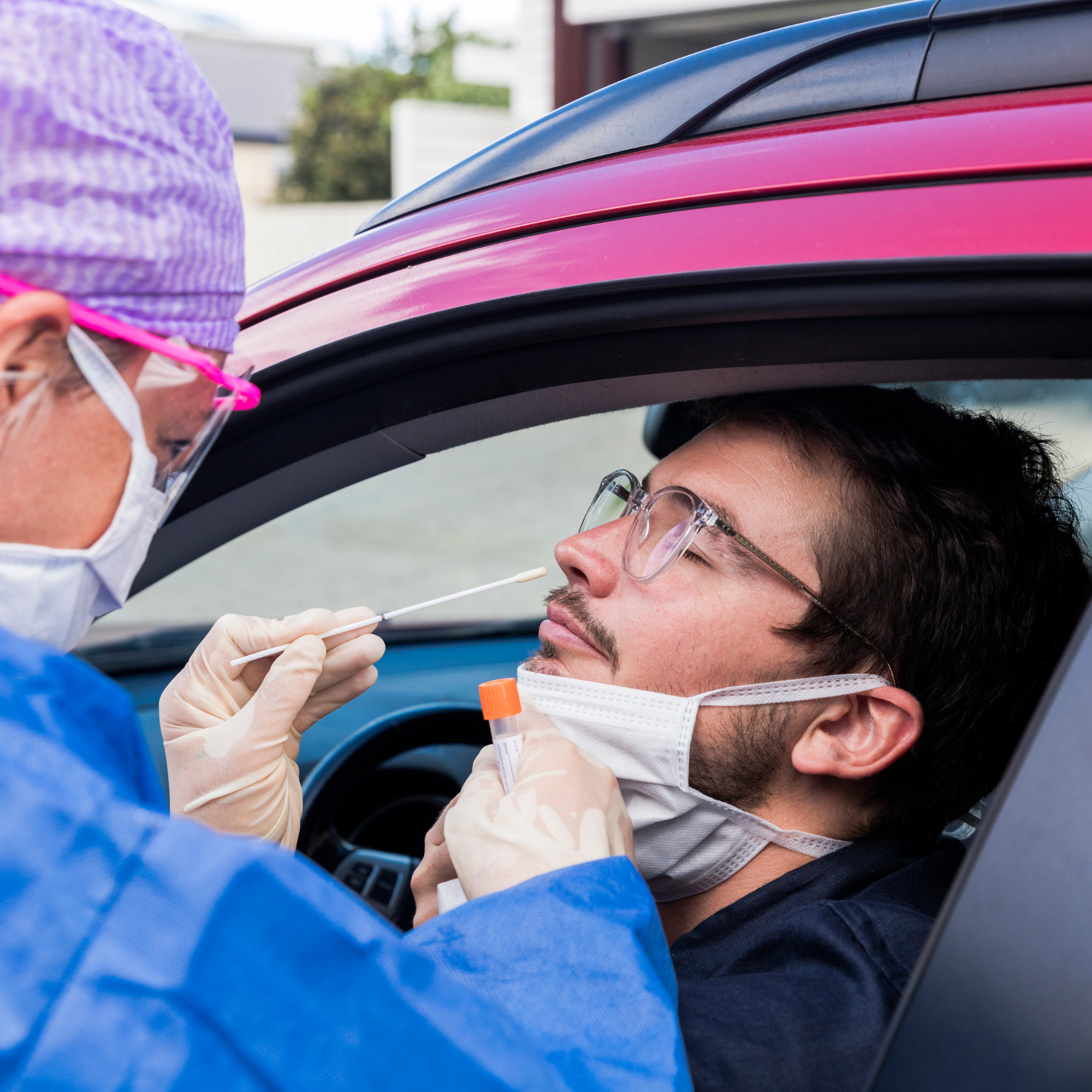 A man in a car undergoing a nasal swab being performed by a woman in a mask